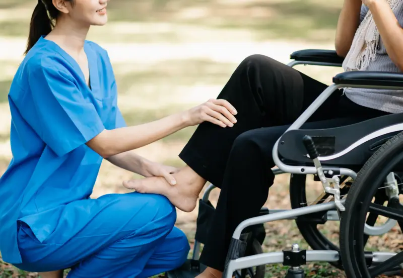Nurse in blue scrubs touching the leg of a patient in a wheel chair.