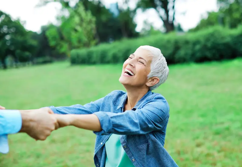 Senior woman laughing outdoors, holding hands with another person.